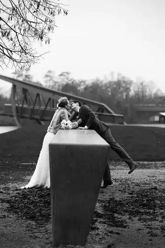 Janina und Florian küssen sich auf einer Betonkonstruktion im Freien, den Blumenstrauß in der Nähe. Eine Brücke und kahle Bäume umrahmen diesen Schwarz-Weiß-Moment, der von Hochzeitsfotograf Ladenburg aufgenommen wurde und zeitlose Romantik in der Nähe der Schweiz heraufbeschwört.