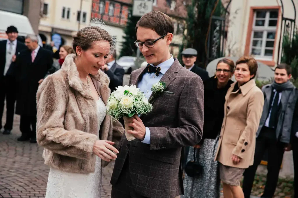Onelovephoto Eine Braut in einer Pelzjacke und ein Bräutigam mit Brille und kariertem Anzug halten gemeinsam einen Blumenstrauß und stehen im Freien vor einer Gruppe lächelnder, warm gekleideter Menschen im Hintergrund. Hochzeitsfotografie in Mannheim und Heidelberg