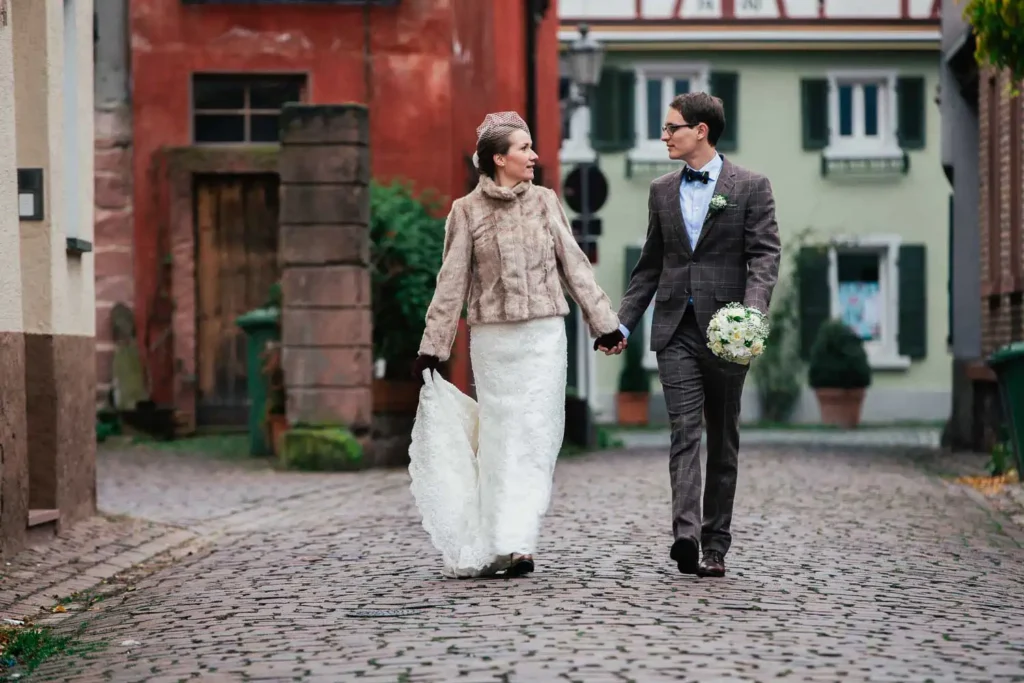 Onelovephoto Eine Braut in einem weißen Kleid und einer Pelzjacke geht Hand in Hand mit einem Bräutigam in einem braunen Anzug, der einen Blumenstrauß hält. Sie schlendern eine von bunten Häusern gesäumte Kopfsteinpflasterstraße entlang, schauen sich an und lächeln. Hochzeitsfotografie in Mannheim und Heidelberg