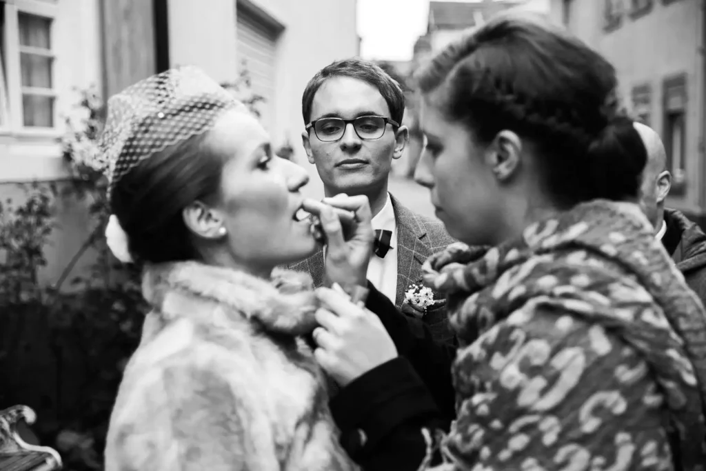 Onelovephoto Eine Frau trägt einer anderen Frau mit Pelzmantel und Hut Lippenstift auf, während ein Mann mit Brille und Anzug hinter ihnen steht und sie beobachtet. Die Szene spielt sich im Freien in einer engen Straße ab. Hochzeitsfotografie in Mannheim und Heidelberg