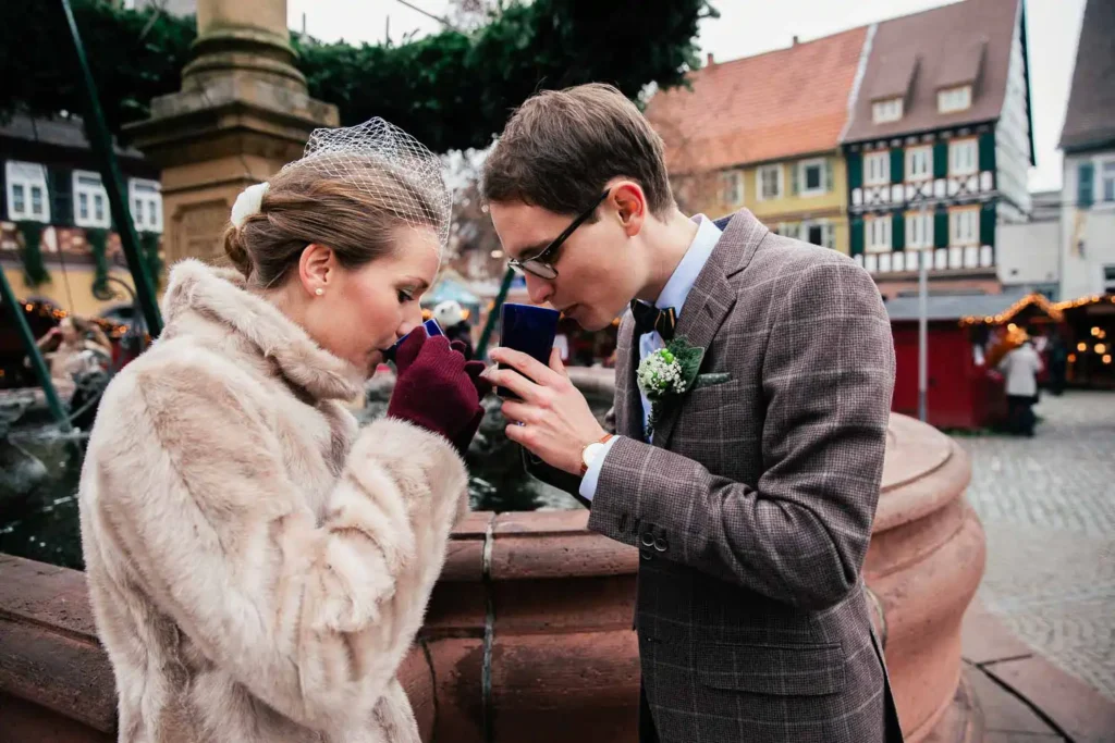 Eine Braut im Pelzmantel und ein Bräutigam im Anzug trinken gemeinsam aus Bechern auf einem Markt im Freien, stehen an einem Steinbrunnen mit festlichen Gebäuden und Lichtern im Hintergrund - Momente, die ich als Hochzeitsfotograf in Ladenburg gerne festhalte. Onelovephoto - Hochzeitsfotografie in Mannheim und Heidelberg