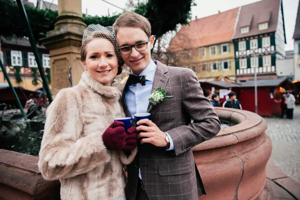 Ein lächelndes, formell gekleidetes Paar - sie im Pelzmantel und Handschuhen, er im Anzug mit Fliege und Ansteckblume - steht Arm in Arm mit Tassen an einem Brunnen auf einem Markt im Freien. Als Hochzeitsfotograf in Ladenburg halte ich solche Momente fest. Onelovephoto - Hochzeitsfotografie in Mannheim und Heidelberg