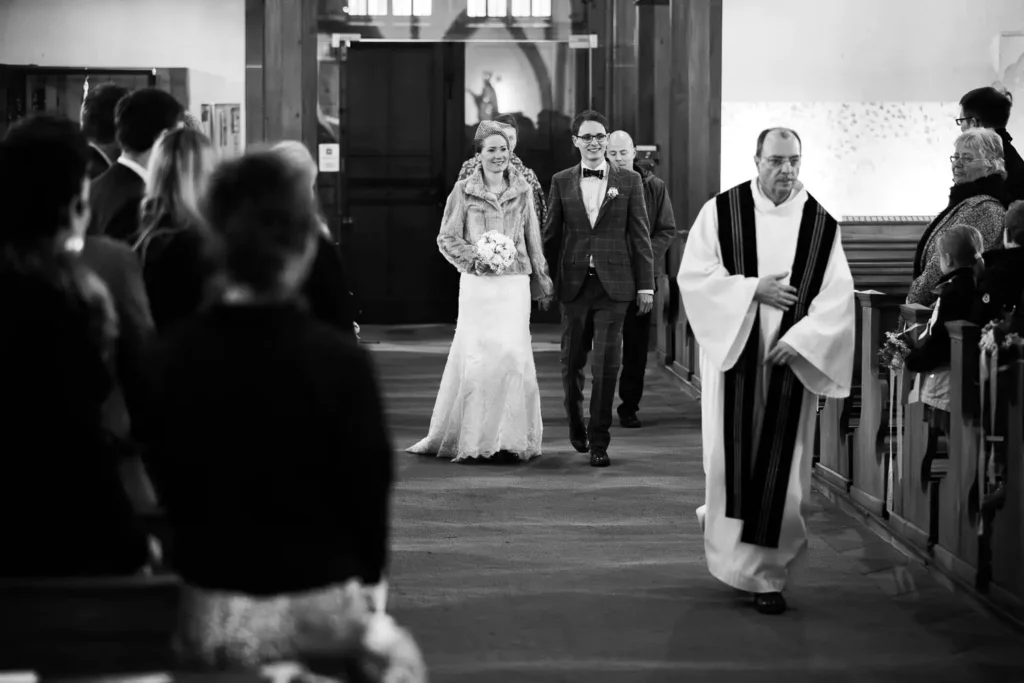 Onelovephoto Eine Braut und ein Bräutigam schreiten gemeinsam lächelnd den Gang in einer Kirche hinunter, während ein Priester sie anführt. Die Gäste sitzen auf beiden Seiten und beobachten das Paar. Die Szene ist in schwarz-weiß gehalten. Hochzeitsfotografie in Mannheim und Heidelberg