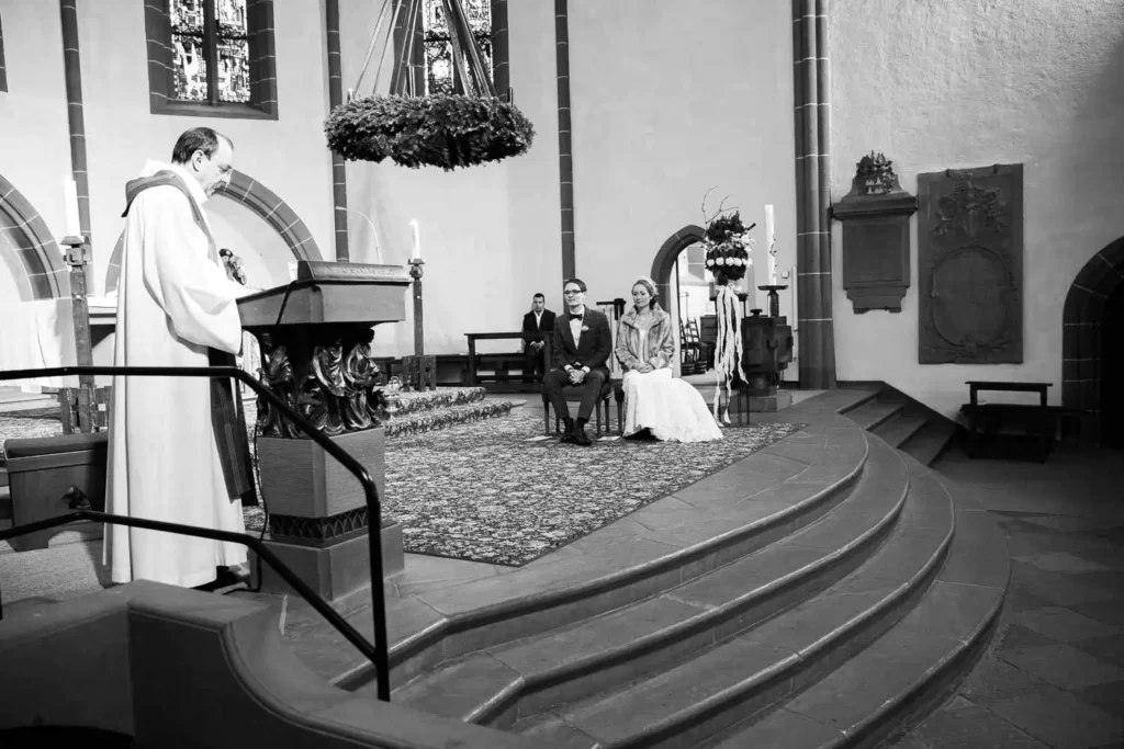 Onelovephoto Eine Braut und ein Bräutigam sitzen während einer Hochzeitszeremonie in einer Kirche gemeinsam am Altar, während ein Priester am Lesepult steht und liest. Die Kulisse besteht aus gewölbten Fenstern und prunkvollen Dekorationen. Hochzeitsfotografie in Mannheim und Heidelberg
