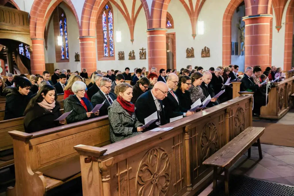 Onelovephoto Eine Gemeinde von Menschen, die in Holzbänken in einer Kirche sitzen und stehen, Papiere in der Hand halten und daraus lesen, mit Bogenfenstern und Glasmalereien im Hintergrund. Hochzeitsfotografie in Mannheim und Heidelberg