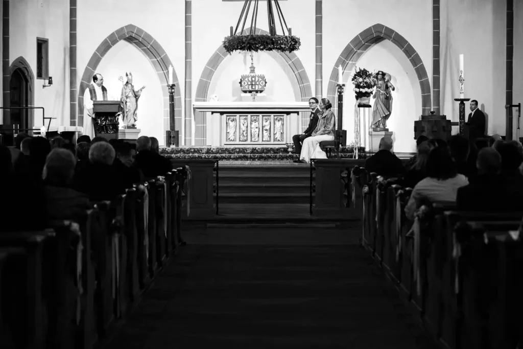 Onelovephoto Braut und Bräutigam sitzen während einer Hochzeitszeremonie am Altar einer Kirche und blicken auf einen Priester, der von einem Rednerpult aus spricht. Die Gäste sitzen in den Kirchenbänken und verfolgen die Zeremonie. Das Foto ist in schwarz-weiß gehalten. Hochzeitsfotografie in Mannheim und Heidelberg