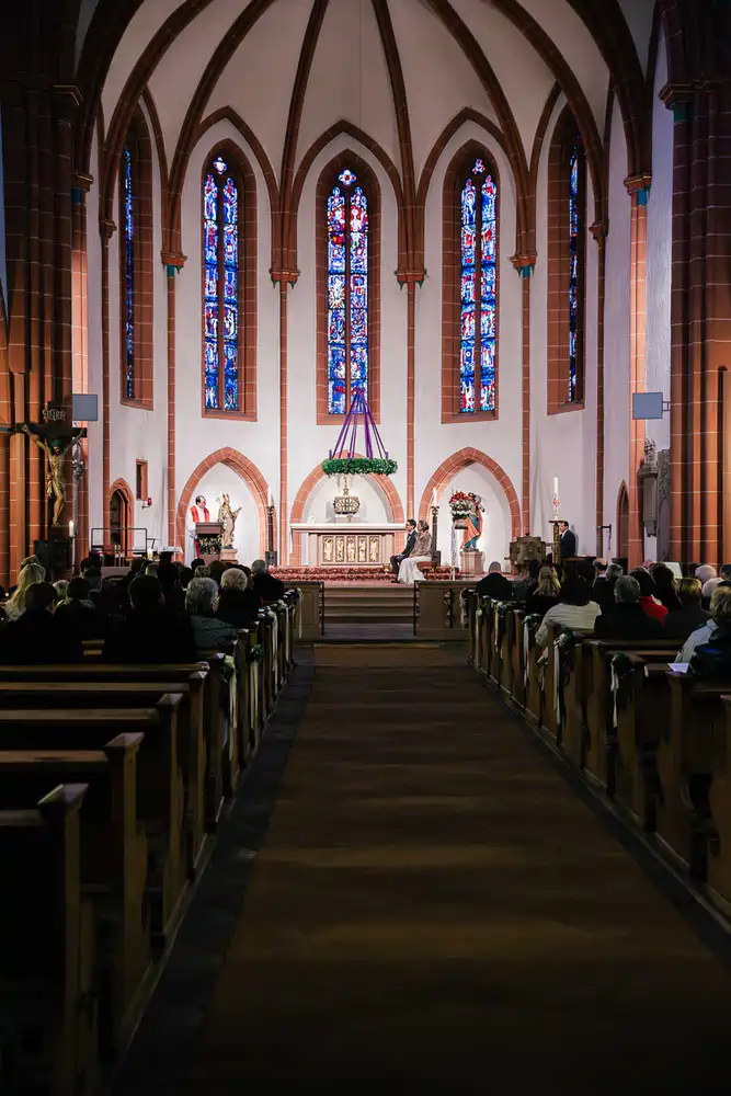 Onelovephoto Blick in den Mittelgang einer Kirche mit hohen Gewölbedecken, Buntglasfenstern und voll besetzten Kirchenbänken, die dem Altar zugewandt sind. Hochzeitsfotografie in Mannheim und Heidelberg