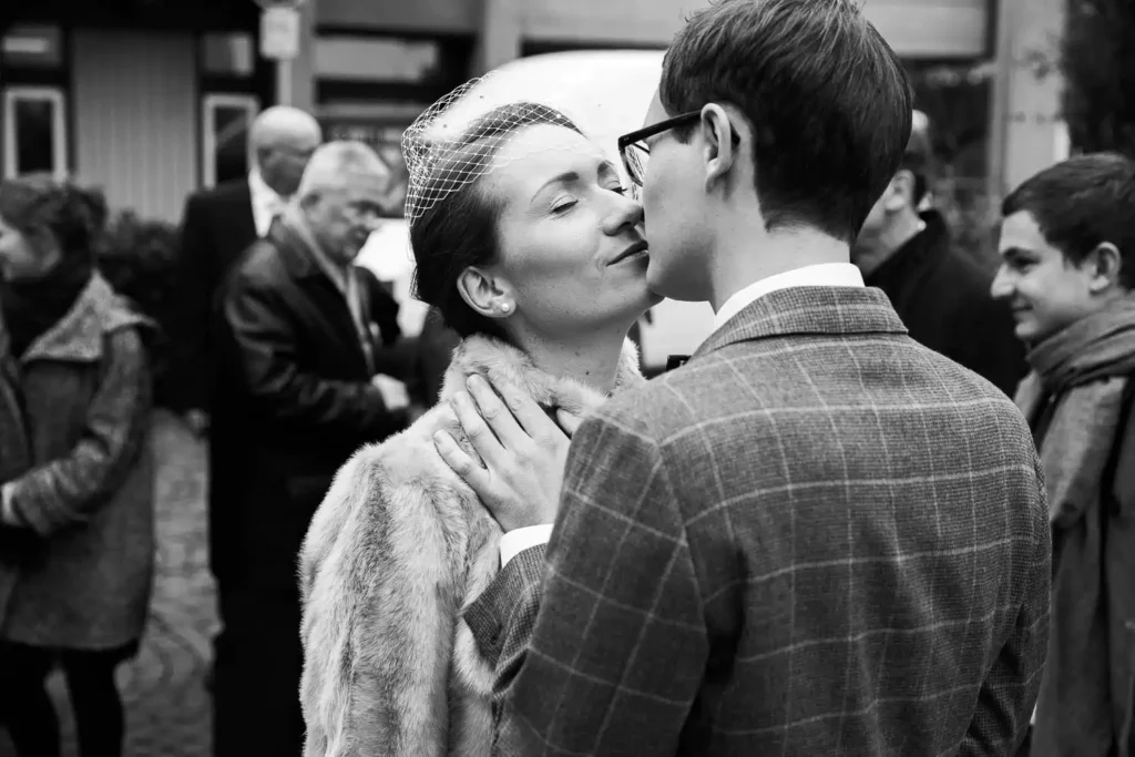 Onelovephoto Ein Paar in Vintage-Kleidung teilt einen intimen Moment und küsst sich fast, umgeben von Menschen im Freien. Die Frau trägt einen Pelzmantel und einen Fascinator, während der Mann eine Brille und einen karierten Anzug trägt. Das Bild ist in schwarz-weiß gehalten. Hochzeitsfotografie in Mannheim und Heidelberg