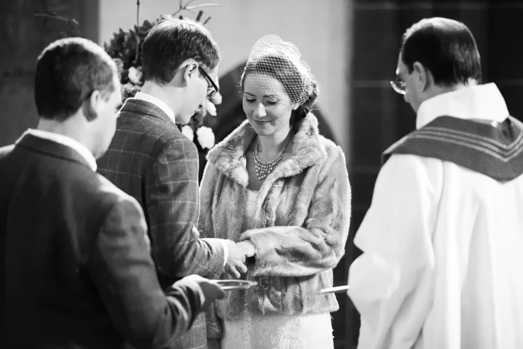 Onelovephoto Eine Braut und ein Bräutigam tauschen während einer Hochzeitszeremonie die Ringe aus, wobei die Braut lächelt. Zwei Männer, einer mit einem Tablett in der Hand und einer in kirchlicher Kleidung, stehen in der Nähe. Das Bild ist in schwarz-weiß gehalten. Hochzeitsfotografie in Mannheim und Heidelberg