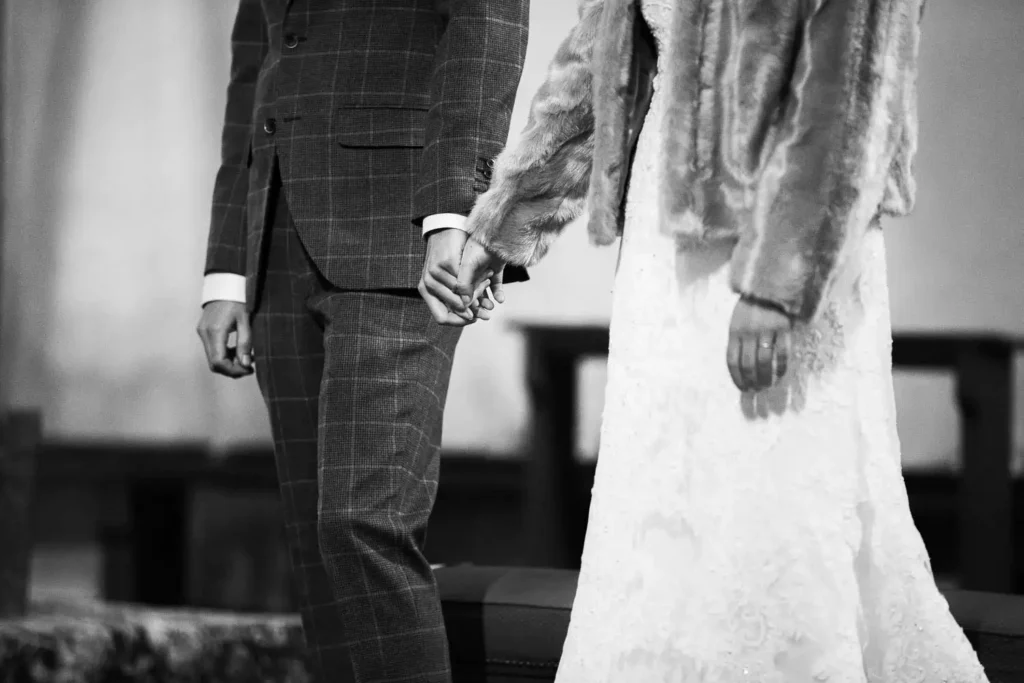Onelovephoto Schwarz-Weiß-Foto eines Paares, das sich an den Händen hält; eine Person trägt einen karierten Anzug, die andere ein Spitzenkleid mit einem Pelzmantel, was auf eine Hochzeit oder eine formelle Veranstaltung hindeutet. Ihre Gesichter sind nicht zu erkennen. Hochzeitsfotografie in Mannheim und Heidelberg