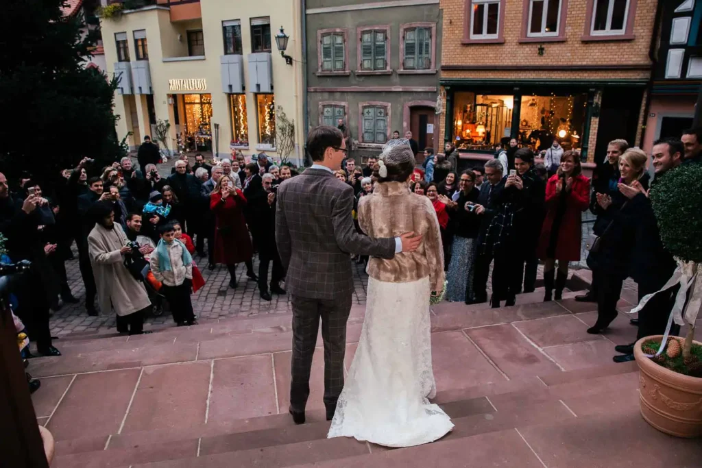 Onelovephoto Ein frisch verheiratetes Paar steht auf steinernen Stufen vor einer Menge gut gekleideter Gäste auf einem charmanten Stadtplatz, während die Leute Fotos machen und ihre Hochzeit feiern. Hochzeitsfotografie in Mannheim und Heidelberg