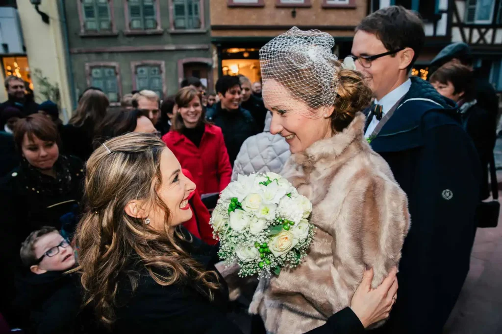 Eine Braut in Pelzmantel und Schleier lächelt, während sie einen Strauß weißer Blumen in der Hand hält. Sie begrüßt eine fröhliche Frau bei einem Treffen im Freien, umgeben von lächelnden Gästen - festliche Momente Als Hochzeitsfotograf in Ladenburg eingefangen. Onelovephoto - Hochzeitsfotografie in Mannheim und Heidelberg