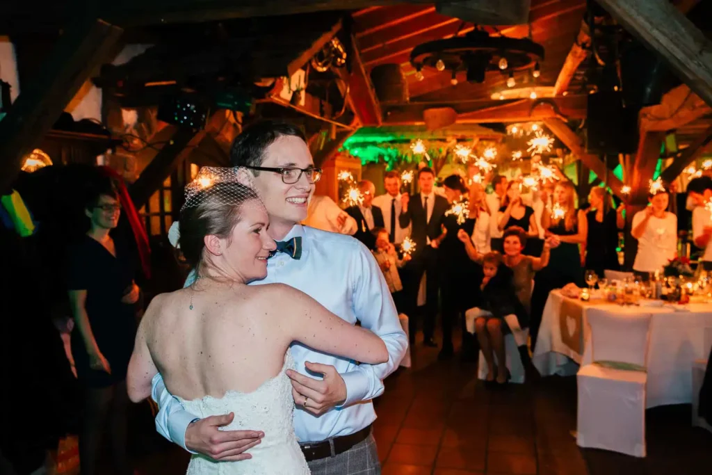 Onelovephoto Braut und Bräutigam tanzen auf ihrer Hochzeitsfeier und lächeln, während die Gäste im Hintergrund mit Wunderkerzen in einem warm beleuchteten, rustikalen Lokal feiern. Hochzeitsfotografie in Mannheim und Heidelberg