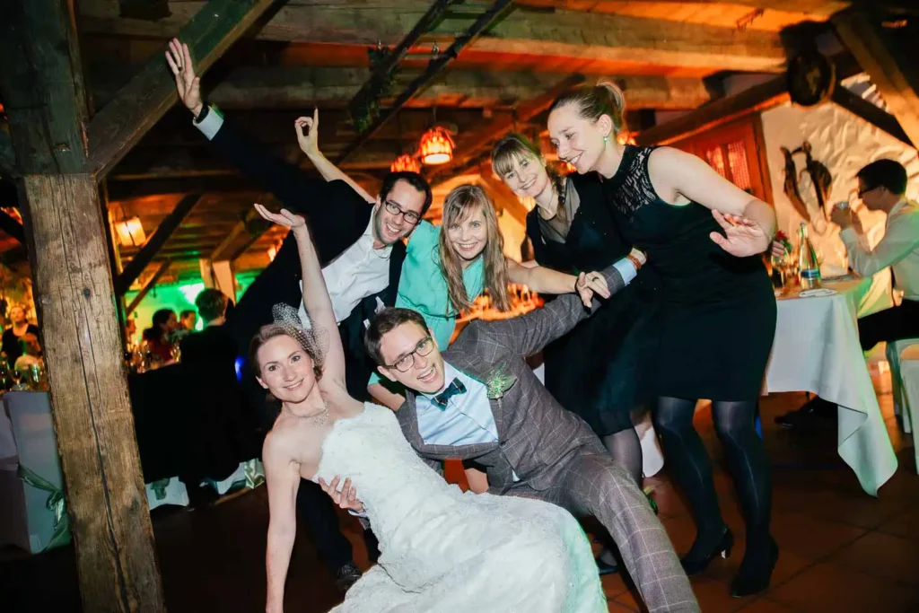 Braut und Bräutigam posieren spielerisch mit vier Gästen auf ihrer Hochzeitsfeier. Als Hochzeitsfotograf in Ladenburg habe ich sie alle lächelnd und angelehnt unter Holzbalken eingefangen, was eine lustige und fröhliche Atmosphäre schafft. Onelovephoto - Hochzeitsfotografie in Mannheim und Heidelberg