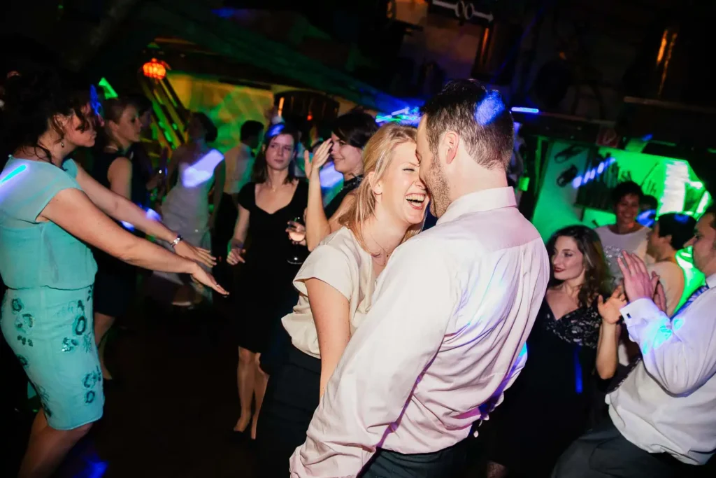 Onelovephoto Eine Gruppe von Menschen tanzt und lacht in einer lebhaften, farbenfrohen Partyumgebung. Zwei Personen in der Mitte, die sich eng gegenüberstehen, lächeln und wirken fröhlich, umgeben von anderen, die sich bewegen und die energiegeladene Atmosphäre genießen. Hochzeitsfotografie in Mannheim und Heidelberg