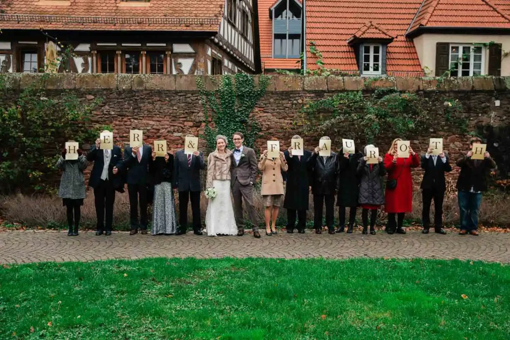 Als Hochzeitsfotograf in Ladenburg habe ich dieses fröhliche Wedding-Shooting festgehalten: Die Hochzeitsgesellschaft steht im Freien, hält gemeinsam Buchstaben hoch und bildet den Schriftzug "Herr & Frau Roth". Onelovephoto - Hochzeitsfotografie in Mannheim und Heidelberg