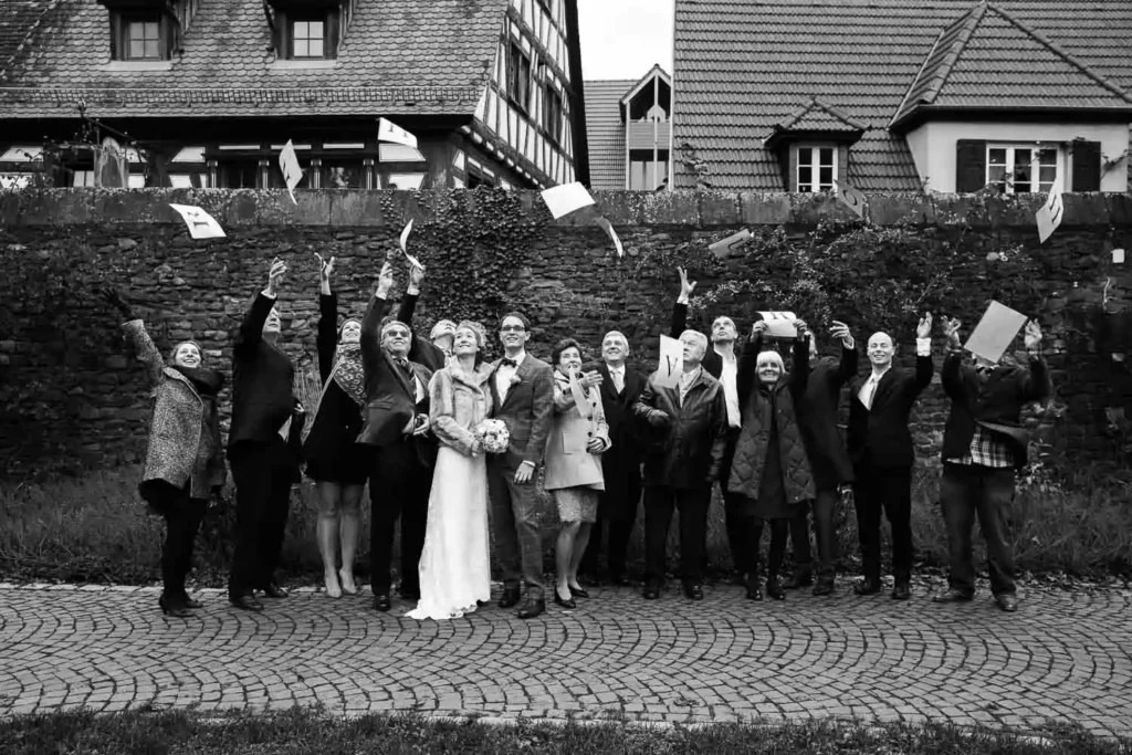 Onelovephoto Eine Gruppe von Menschen, darunter Braut und Bräutigam, steht im Freien vor einer Steinmauer und Häusern und wirft freudig Papiere in die Luft. Das Foto ist schwarz-weiß, und alle scheinen zu feiern. Hochzeitsfotografie in Mannheim und Heidelberg