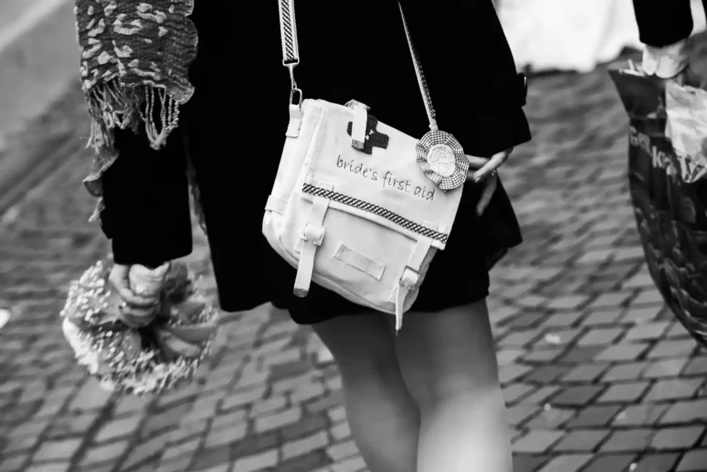 Onelovephoto Eine Person, die Blumen in der Hand hält und eine weiße Tasche mit der Aufschrift "Erste Hilfe für die Braut" trägt, geht auf einer Straße mit Kopfsteinpflaster; das Bild ist in schwarz-weiß gehalten. Hochzeitsfotografie in Mannheim und Heidelberg
