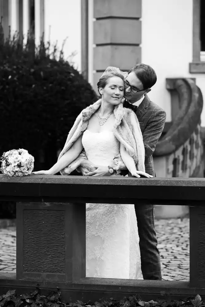 Onelovephoto Eine Braut im Spitzenkleid und Pelzmantel steht vor einem Bräutigam im Anzug und mit Brille. Sie lächeln sich auf einer Kopfsteinpflasterstraße liebevoll an, während ein Blumenstrauß auf dem Geländer neben ihnen ruht. Hochzeitsfotografie in Mannheim und Heidelberg