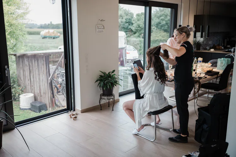 Onelovephoto Eine Frau in einem weißen Gewand sitzt auf einem Stuhl, während eine andere Frau ihr Haar in einem Haus frisiert; große Fenster geben den Blick auf eine grüne Außenlandschaft frei, und im Hintergrund ist ein Tisch mit Gegenständen zu sehen. Hochzeitsfotografie in Mannheim und Heidelberg