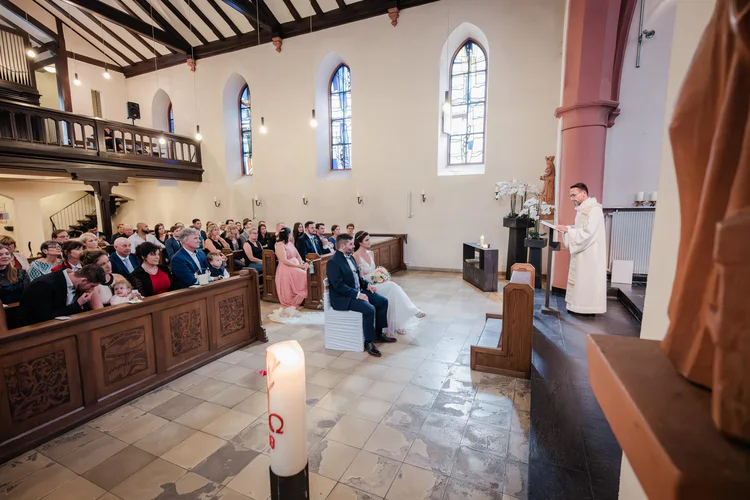 Onelovephoto Eine Hochzeitszeremonie in einer Kirche, bei der die Gäste in den Kirchenbänken sitzen, die Braut und der Bräutigam vorne sitzen und ein Priester am Altar steht und spricht. Buntglasfenster erhellen den Raum. Hochzeitsfotografie in Mannheim und Heidelberg