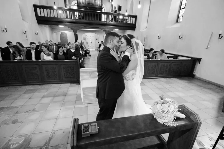 Onelovephoto Eine Braut und ein Bräutigam küssen sich während ihrer Hochzeitszeremonie in einer Kirche am Altar, während die Gäste im Hintergrund sitzen und zusehen. Ein Blumenstrauß liegt auf einem Tisch in der Nähe. Das Bild ist in schwarz-weiß gehalten. Hochzeitsfotografie in Mannheim und Heidelberg