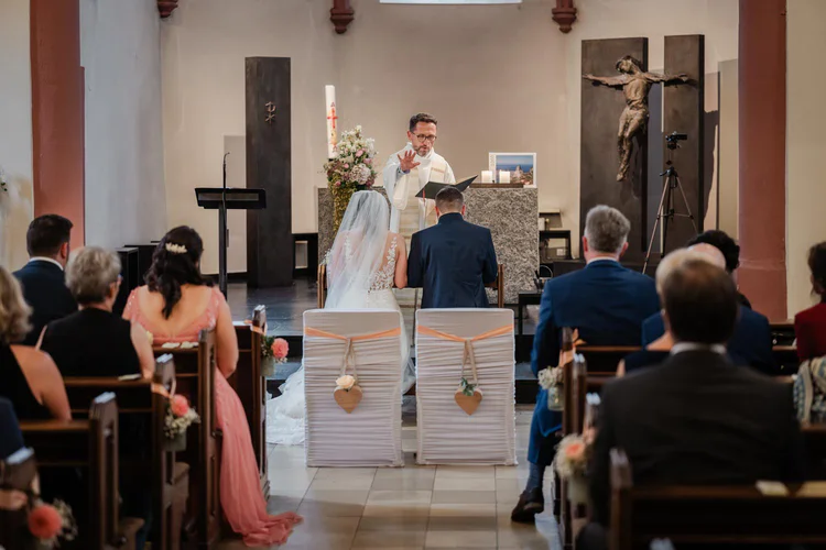 Onelovephoto Braut und Bräutigam sitzen während ihrer Trauung in einer Kirche vor einem Priester am Altar, während die Gäste in den Kirchenbänken sitzen und zusehen. Die geschmückten Stühle haben herzförmige Ornamente auf der Rückseite. Hochzeitsfotografie in Mannheim und Heidelberg