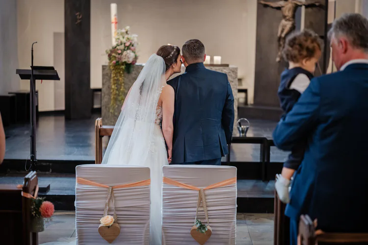 Onelovephoto Braut und Bräutigam sitzen während ihrer Trauung eng beieinander vor einer Kirche, mit dem Rücken zur Kamera. Die Gäste sitzen in der Nähe, und im Hintergrund sind der Altar und der Blumenschmuck zu sehen. Hochzeitsfotografie in Mannheim und Heidelberg