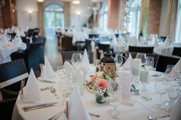 Onelovephoto Eleganter Bankettsaal mit runden Tischen, die für eine Veranstaltung gedeckt sind, mit weißen Tischtüchern, gefalteten Servietten, Glaswaren, Kerzen, Blumen und einem dekorativen Laternen-Mittelstück. Sanftes natürliches Licht erfüllt den Raum. Hochzeitsfotografie in Mannheim und Heidelberg