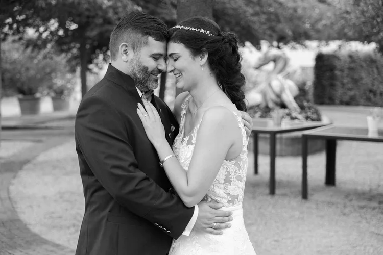 Onelovephoto Eine Braut und ein Bräutigam umarmen sich im Freien und berühren sich lächelnd an der Stirn. Die Braut trägt ein Spitzenkleid und der Bräutigam einen Anzug. Im Hintergrund sind Bäume und eine verschwommene Statue zu sehen. Das Bild ist in schwarz-weiß gehalten. Hochzeitsfotografie in Mannheim und Heidelberg