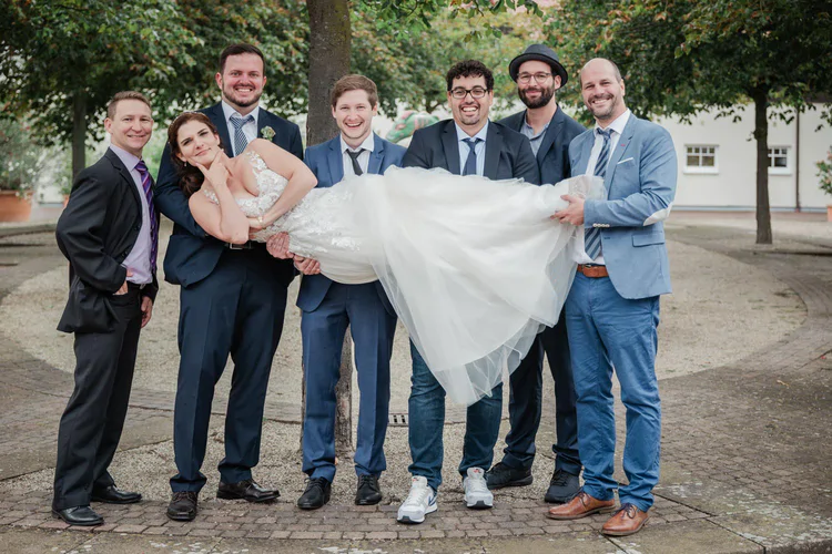 Onelovephoto Eine Braut in einem weißen Kleid wird von sechs lächelnden Männern in Anzügen waagerecht gehalten, die spielerisch im Freien unter Bäumen auf einem gepflasterten Weg posieren. Hochzeitsfotografie in Mannheim und Heidelberg