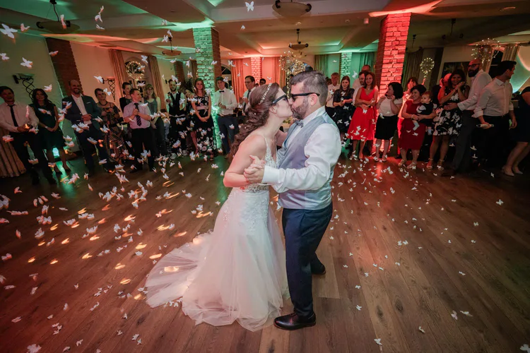 Onelovephoto Braut und Bräutigam tanzen ihren ersten Tanz auf einem Holzboden, umgeben von Gästen und fallendem weißem Konfetti, in einer warm beleuchteten Empfangshalle. Hochzeitsfotografie in Mannheim und Heidelberg