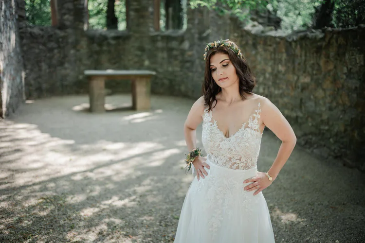 Onelovephoto Eine Braut in einem weißen Spitzenhochzeitskleid steht im Freien, die Hände in die Hüften gestemmt, trägt eine Blumenkrone und blickt nach unten, umgeben von Steinmauern und Sonnenlicht, das durch die Bäume fällt. Hochzeitsfotografie in Mannheim und Heidelberg