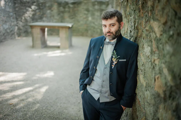 Onelovephoto Ein Mann in einem formellen dunklen Anzug mit einer Boutonniere steht mit den Händen in den Taschen an eine Steinmauer gelehnt im Freien, mit einem unscharfen Steinbauwerk im Hintergrund. Hochzeitsfotografie in Mannheim und Heidelberg