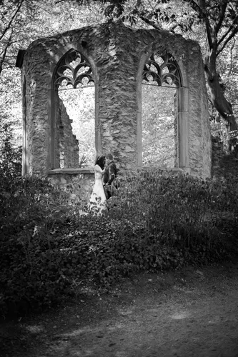 Onelovephoto Ein Paar umarmt sich vor einer steinernen Ruine mit hohen, gewölbten Fenstern, umgeben von dichtem Laub und Bäumen. Das Foto ist in Schwarz-Weiß gehalten und schafft eine romantische und zeitlose Atmosphäre. Hochzeitsfotografie in Mannheim und Heidelberg