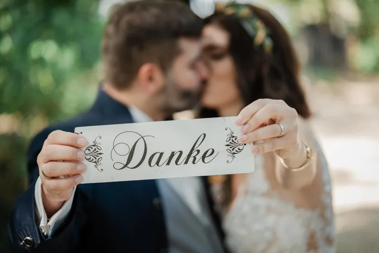 Onelovephoto Ein Paar küsst sich im Hintergrund, während es im Vordergrund ein Schild mit der Aufschrift "Danke" in der Hand hält. Das Bild ist weich fokussiert mit natürlichem Grün hinter ihnen. Hochzeitsfotografie in Mannheim und Heidelberg
