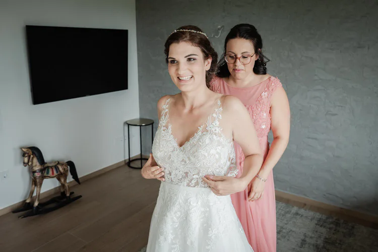 Onelovephoto Eine Braut in einem weißen Spitzenhochzeitskleid lächelt, während eine andere Frau in einem rosa Kleid ihr Kleid in einem modernen Raum mit einem Fernseher an der Wand und einem hölzernen Schaukelpferd im Hintergrund zurechtrückt. Hochzeitsfotografie in Mannheim und Heidelberg