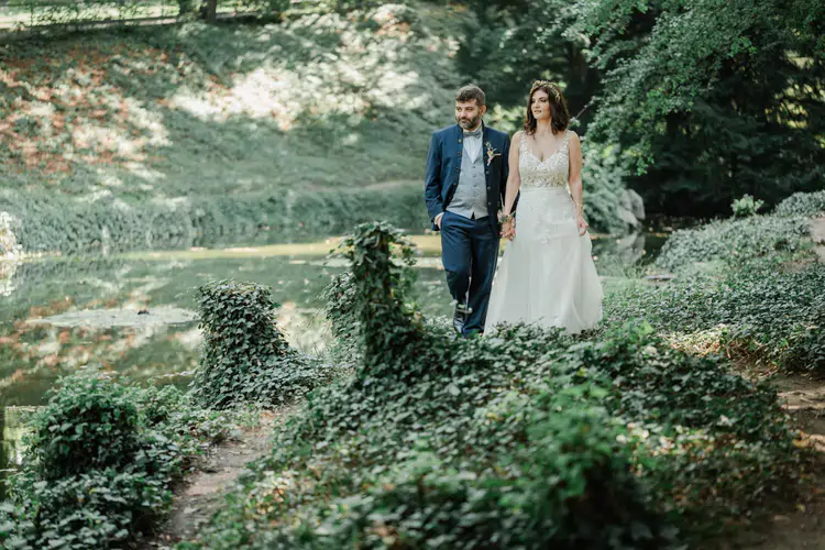 Onelovephoto Braut und Bräutigam spazieren Seite an Seite über einen üppigen, grünen, mit Efeu bewachsenen Weg neben einem ruhigen Teich in einer sonnenbeschienenen Außenanlage. Hochzeitsfotografie in Mannheim und Heidelberg