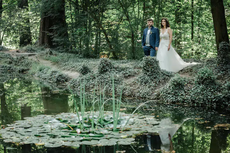 Onelovephoto Ein Brautpaar in Hochzeitskleidung steht an einem üppigen, grünen Teich mit Seerosenblättern, umgeben von dichtem Wald, in dem sich die Bäume und das Paar im stillen Wasser spiegeln. Hochzeitsfotografie in Mannheim und Heidelberg