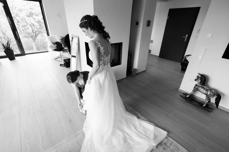 Onelovephoto Eine Braut in einem weißen Hochzeitskleid steht in einem Raum, während eine andere Frau ihr Kleid zurechtrückt. Der Raum hat Holzböden, ein Spielzeug-Schaukelpferd, einen Stuhl und große Fenster, durch die natürliches Licht einfällt. Hochzeitsfotografie in Mannheim und Heidelberg