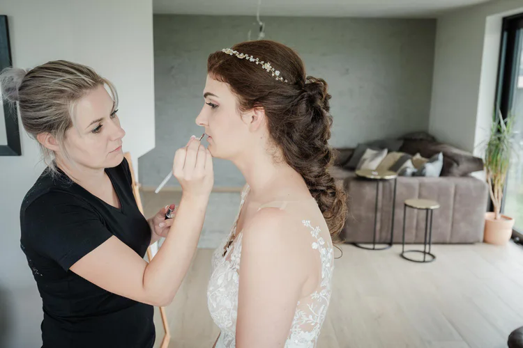 Onelovephoto Eine Visagistin trägt einer Braut mit langen braunen Haaren in einem weißen Kleid Lippenstift auf. Sie steht in einem hellen, modernen Wohnzimmer mit einer Couch und großen Fenstern im Hintergrund. Hochzeitsfotografie in Mannheim und Heidelberg