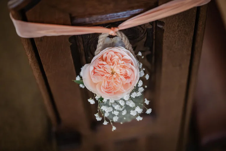 Onelovephoto Eine pfirsichfarbene Rose und weiße Schleierkrautblüten sind mit einem pfirsichfarbenen Band an eine dunkle Holzbank gebunden und bilden eine elegante Blumendekoration. Hochzeitsfotografie in Mannheim und Heidelberg