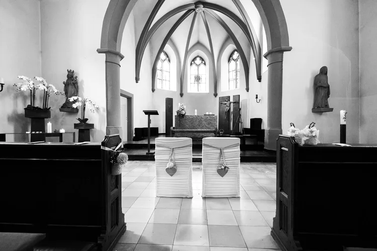 Onelovephoto Schwarz-Weiß-Foto eines Kirchenraums mit zwei mit herzförmigen Ornamenten geschmückten Stühlen vor einem Altar. Zu sehen sind eine gewölbte Decke, Buntglasfenster, Statuen und Blumenarrangements. Hochzeitsfotografie in Mannheim und Heidelberg