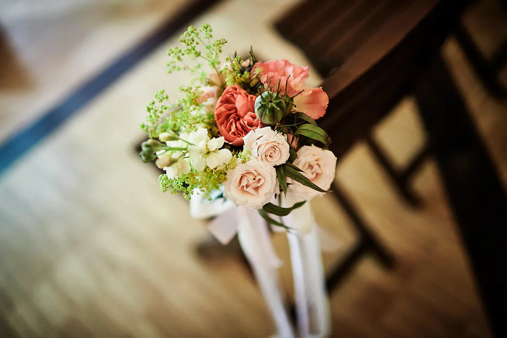 Onelovephoto Eine Nahaufnahme eines Blumenstraußes mit rosa, weißen und pfirsichfarbenen Rosen, grünem Laub und weißem Band, der auf einem Stuhl in einem sanft beleuchteten Innenraum ruht. Hochzeitsfotografie in Mannheim und Heidelberg