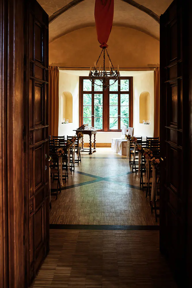 Onelovephoto Blick durch offene Holztüren in einen warm beleuchteten Raum, der für eine Hochzeitszeremonie eingerichtet ist, mit Stuhlreihen gegenüber einem Fenster und einem dekorierten Tisch an der Stirnseite. Ein Kronleuchter hängt von der Decke herab. Hochzeitsfotografie in Mannheim und Heidelberg