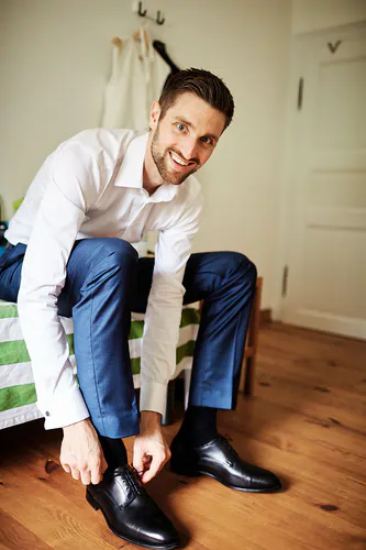 Ein Mann mit kurzen braunen Haaren und einem Bart, der ein weißes Hemd und eine blaue Hose trägt, sitzt auf einem Bett in einem hellen Raum mit Holzboden und lächelt, während er seine schwarzen Schuhe während eines Get-Ready-Shootings in Mittelburg bindet.
