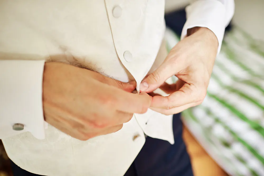 Onelovephoto Nahaufnahme einer Person, die eine cremefarbene Weste über einem weißen Hemd zuknöpft, vor einem verschwommenen Hintergrund mit grünen und weißen Streifen. Hochzeitsfotografie in Mannheim und Heidelberg