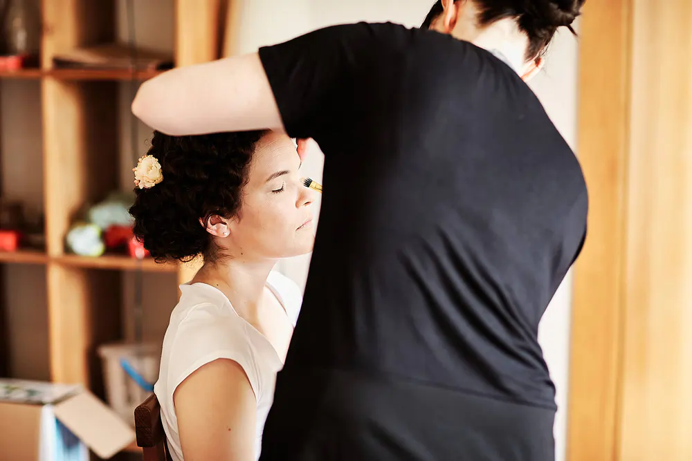 Onelovephoto Eine Frau mit geschlossenen Augen sitzt ruhig da, während eine andere Person ihr Haar frisiert und Blumen anbringt. Die Szenerie ist ein Innenraum mit Regalen im Hintergrund. Hochzeitsfotografie in Mannheim und Heidelberg