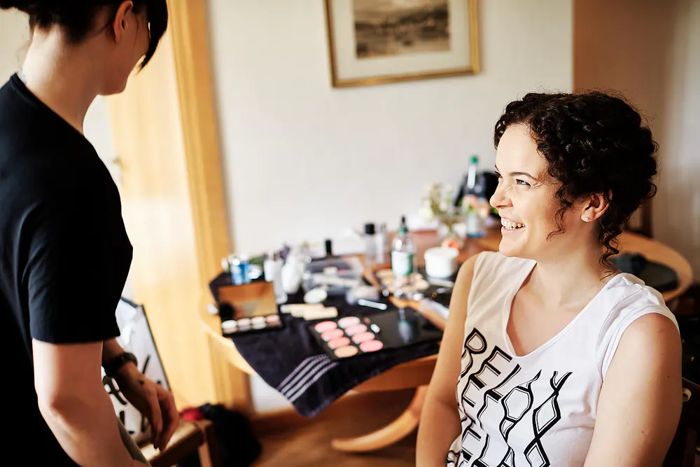 Onelovephoto Eine Frau mit lockigem Haar sitzt lächelnd auf einem Stuhl und trägt ein weißes Hemd, während sich eine andere Person darauf vorbereitet, sie zu schminken. Im Hintergrund ist ein Tisch mit Schminkprodukten und -geräten zu sehen. Hochzeitsfotografie in Mannheim und Heidelberg