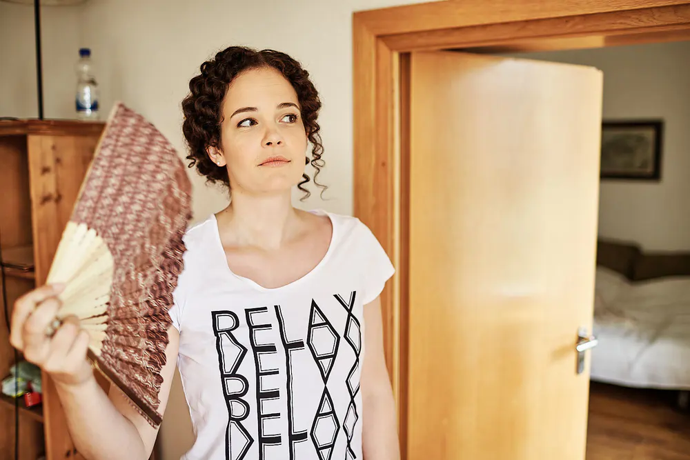 Onelovephoto Eine Frau mit lockigem Haar, die ein weißes T-Shirt mit dem wiederholten Aufdruck "RELAX" trägt, steht in einem Innenraum in der Nähe einer offenen Tür, fächelt sich mit einem Handfächer Luft zu und schaut nachdenklich zur Seite. Hochzeitsfotografie in Mannheim und Heidelberg